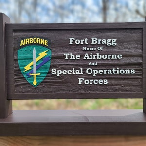 May include: A brown wooden sign with white text that reads "Fort Bragg Home of The Airborne And Special Operations Forces". The sign also features a green, yellow, and purple emblem with a sword and lightning bolts that reads "Airborne".