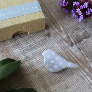 May include: A small, gray bird with white polka dots is on a wooden surface. The bird is made of paper or fabric and has a slightly textured surface.