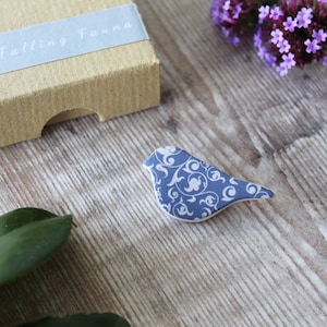 May include: A blue ceramic bird brooch with a white floral pattern. The bird is sitting on a wooden surface.