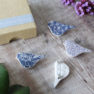 May include: Four ceramic bird brooches with a blue and white damask pattern. The brooches are on a light wood surface. The brooches are in the shape of a bird with a long tail. The brooches are a part of a set of four.