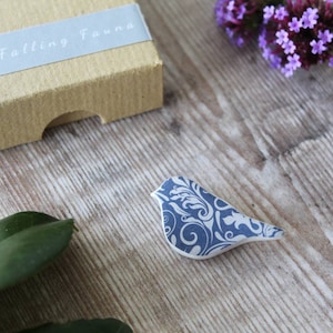 May include: A blue and white ceramic bird brooch with a swirling pattern. The brooch is on a wooden surface.
