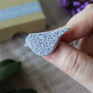 May include: A white ceramic bird brooch with a gray and white floral pattern.