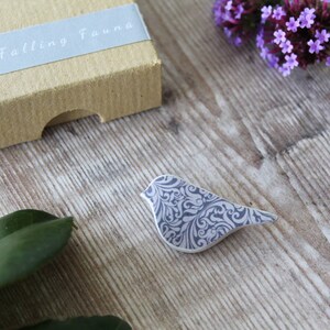 May include: A small, gray ceramic bird brooch with a white and gray floral pattern. The brooch is sitting on a wooden surface.