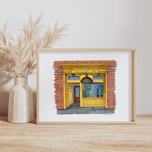 May include: Watercolor painting of a storefront with a yellow facade, red brick walls, and a glass window. The artwork is framed and displayed on a wooden surface. A vase with dried plants is in the background.