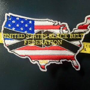 May include: An embroidered patch in the shape of the United States with a black and white striped band across the middle. The band has the text "UNITED STATES BLACK BELT FEDERATION" in yellow lettering. The patch is on a dark gray background.