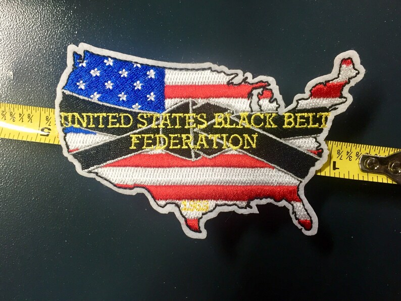 May include: An embroidered patch in the shape of the United States with a black and white striped band across the middle. The band has the text "UNITED STATES BLACK BELT FEDERATION" in yellow lettering. The patch is on a dark gray background.