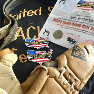 May include: A black martial arts uniform with a gold embroidered patch that reads "United States Black Belt Federation".  Two smaller patches with the same text and a US map design are also visible. A brown leather belt, a pair of brown leather boxing gloves, and a brown leather kickboxing shin guard are also in the image. A certificate of membership in the United States Black Belt Federation is visible in the background.