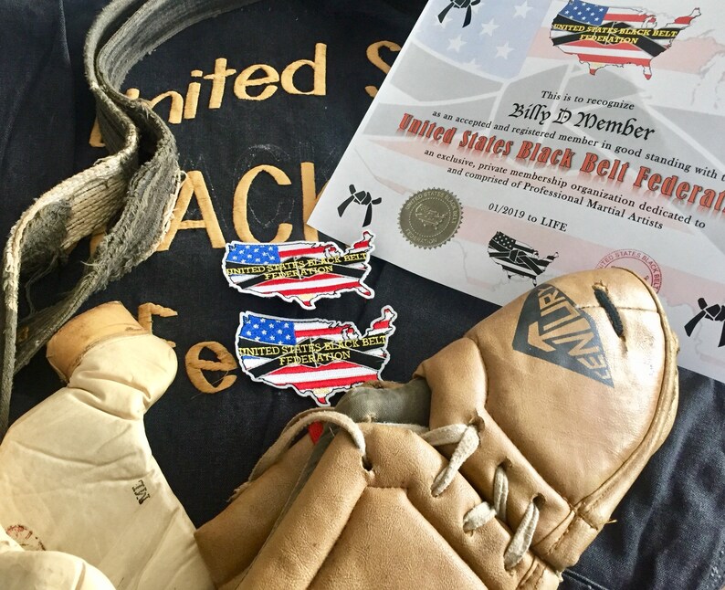 May include: A black martial arts uniform with a gold embroidered patch that reads "United States Black Belt Federation".  Two smaller patches with the same text and a US map design are also visible. A brown leather belt, a pair of brown leather boxing gloves, and a brown leather kickboxing shin guard are also in the image. A certificate of membership in the United States Black Belt Federation is visible in the background.