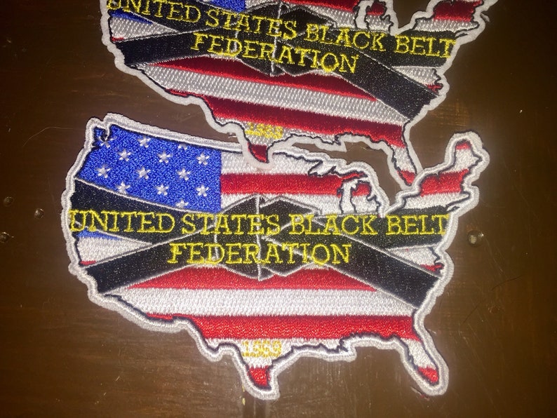 May include: A red, white, and blue embroidered patch shaped like the United States of America. The patch has a black belt across it with the text "UNITED STATES BLACK BELT FEDERATION" in yellow letters.