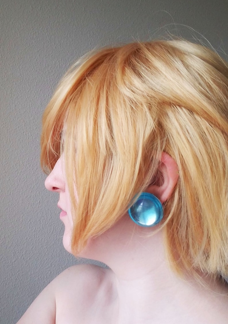 Op de afbeelding: Zijaanzicht van een persoon met lichtblond haar die grote, ronde, doorschijnende blauwe oorbellen draagt. De oorbellen zijn een opvallend accessoire. De achtergrond is een effen, lichtgrijze muur.