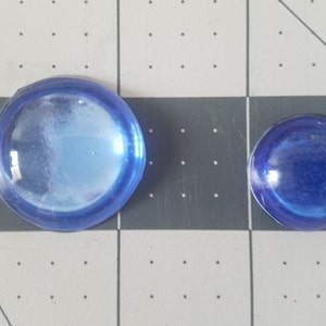 Op de afbeelding: Twee blauwe glazen cabochons van verschillende formaten. De grotere heeft een diameter van ongeveer 3,8 cm, terwijl de kleinere ongeveer 2,5 cm meet. Beide zijn rond met een licht gewelfd oppervlak en een doorschijnend uiterlijk.