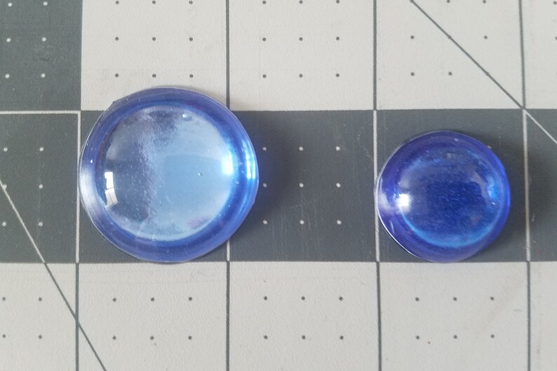 Op de afbeelding: Twee blauwe glazen cabochons van verschillende formaten. De grotere heeft een diameter van ongeveer 3,8 cm, terwijl de kleinere ongeveer 2,5 cm meet. Beide zijn rond met een licht gewelfd oppervlak en een doorschijnend uiterlijk.