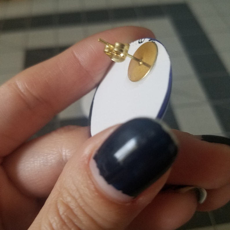 Op de afbeelding: Close-up van een ovale oorbel met een goudkleurige pin en achterkant. De oorbel heeft een wit oppervlak met een blauwe rand. De oorbel wordt door een persoon vastgehouden.