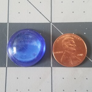 Op de afbeelding: Een ronde, doorschijnende blauwe glazen edelsteen naast een koperen cent. De edelsteen is glad en heeft een enigszins onregelmatige vorm. De cent toont het profiel van een man en het jaar 2015.