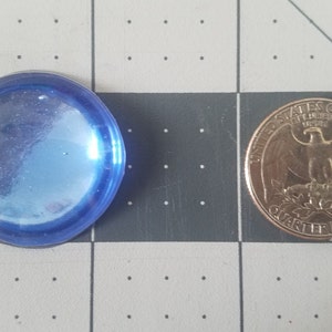 Op de afbeelding: Een helder, blauw, rond glazen object naast een Amerikaanse kwart dollar. Het glazen object heeft een licht iriserende kwaliteit. De munt toont de afbeelding van een adelaar en de woorden "United States of America".