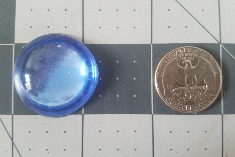 Op de afbeelding: Een helder, blauw, rond glazen object naast een Amerikaanse kwart dollar. Het glazen object heeft een licht iriserende kwaliteit. De munt toont de afbeelding van een adelaar en de woorden "United States of America".