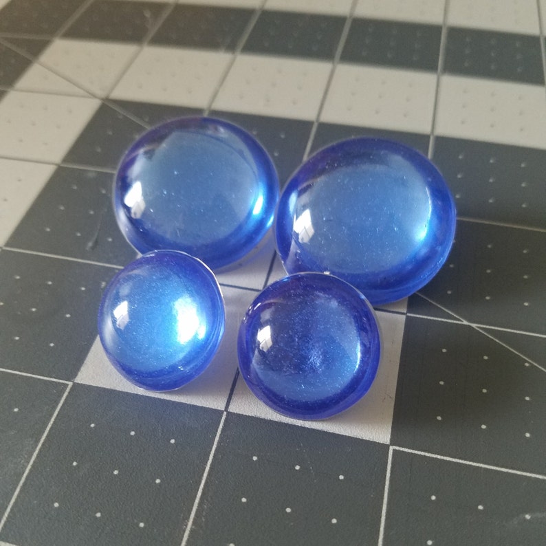 Op de afbeelding: Vier blauwe decoratieve glazen stenen van verschillende formaten. De stenen hebben een afgeronde vorm en een glanzende afwerking, die het licht reflecteert. De stenen zijn gerangschikt op een grijs en wit geblokt oppervlak.