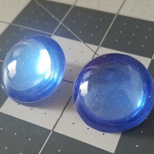 Op de afbeelding: Twee ronde, doorschijnende blauwe glazen cabochons. De cabochons hebben een gewelfde vorm en een glanzende afwerking, die licht reflecteert. Ze liggen op een grijs en zwart geruit patroon.