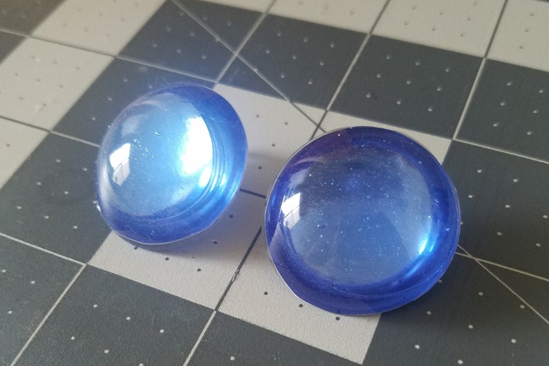 Op de afbeelding: Twee ronde, doorschijnende blauwe glazen cabochons. De cabochons hebben een gewelfde vorm en een glanzende afwerking, die licht reflecteert. Ze liggen op een grijs en zwart geruit patroon.