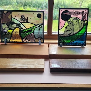 May include: Two stained glass panels displayed on wooden stands. The first panel features a rural scene with a house, trees, a fox, a badger, and an owl. The second panel shows a cat and an owl in a boat with the text "Sail Away".