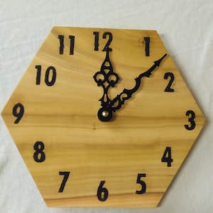May include: A wooden, hexagonal clock with black numerals and hands. The clock face is a light brown color, and the hands are positioned to show the time. The clock is a decorative item.