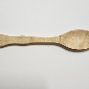May include: A wooden spoon with a light brown finish. The spoon has a rounded bowl and a handle with a slightly curved design. The spoon is approximately 30 cm long, suitable for cooking and serving.