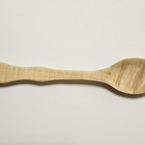 May include: A light brown wooden spoon. The spoon features a rounded bowl and a gently curved handle. The wood grain is visible, adding texture to the utensil. The spoon is approximately 20 cm long.