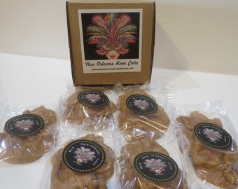 May include: A box of New Orleans Rum Cake with a label featuring a mask design. Several individually wrapped cakes are visible, each with a round sticker. The cakes are light brown and appear to be a baked good.