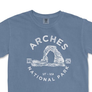 May include: A blue t-shirt featuring a white graphic of the Arches National Park in Utah. The design includes the words "Arches National Park" and "UT - USA" with the establishment date of 1929. The shirt is a short-sleeved crew neck.