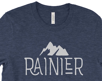 Peut inclure: Un t-shirt bleu marine avec le mot "RAINIER" en gris clair, surmonté d'un motif de montagne. Le t-shirt est fait d'une matière douce.