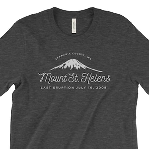Puede incluir: Camiseta gris oscuro con un gráfico blanco del Monte Santa Helena, un volcán en el condado de Skamania, Washington. El gráfico incluye el texto "Mount St. Helens" y "Last Eruption July 10, 2008".