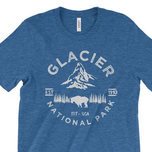 Peut inclure: Un t-shirt bleu avec un motif graphique blanc du logo du parc national de Glacier. Le logo repr&eacute;sente une cha&icirc;ne de montagnes, un bison et le texte "Glacier National Park Est. 1910 MT-USA".
