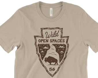 May include: A tan t-shirt with a brown graphic of a vintage park ranger badge. The badge features a bison silhouette, the text "Wild Open Spaces", and the number "56".