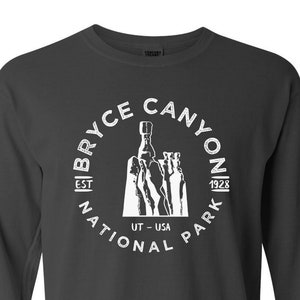 May include: A dark gray long-sleeve t-shirt with a white graphic of Bryce Canyon National Park, Utah, USA. The graphic includes the park's name, the year it was established (1928), and a silhouette of the iconic hoodoos.