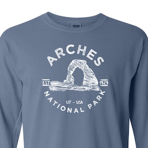 May include: A blue long-sleeve t-shirt with a white graphic of the Arches National Park logo. The logo features a drawing of Delicate Arch, the park's iconic natural sandstone arch, with the words "Arches National Park" and "Est. 1929" around it.