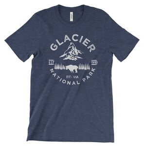 Peut inclure: Un t-shirt bleu marine avec un motif blanc du parc national de Glacier. Le motif comprend le nom du parc, une cha&icirc;ne de montagnes, un bison et le texte "Est. 1910, MT-USA, National Park".