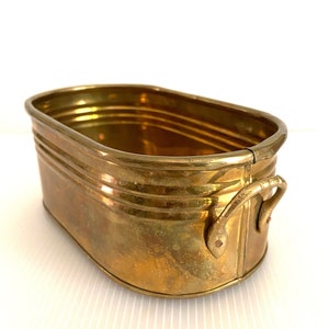 Puede incluir: Un pequeño macetero ovalado de latón con dos asas. El macetero tiene un acabado ligeramente desgastado y es perfecto para añadir un toque de encanto vintage a la decoración de tu hogar.