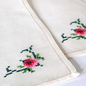 K&ouml;nnte beinhalten: Off-white Leinen-Servietten mit gestickten Blumenmustern. Jede Serviette hat eine kreuzgestickte rosa Blume mit gr&uuml;nen Bl&auml;ttern und Stielen in der Ecke. Die Kanten sind mit einem dekorativen Stich versehen.