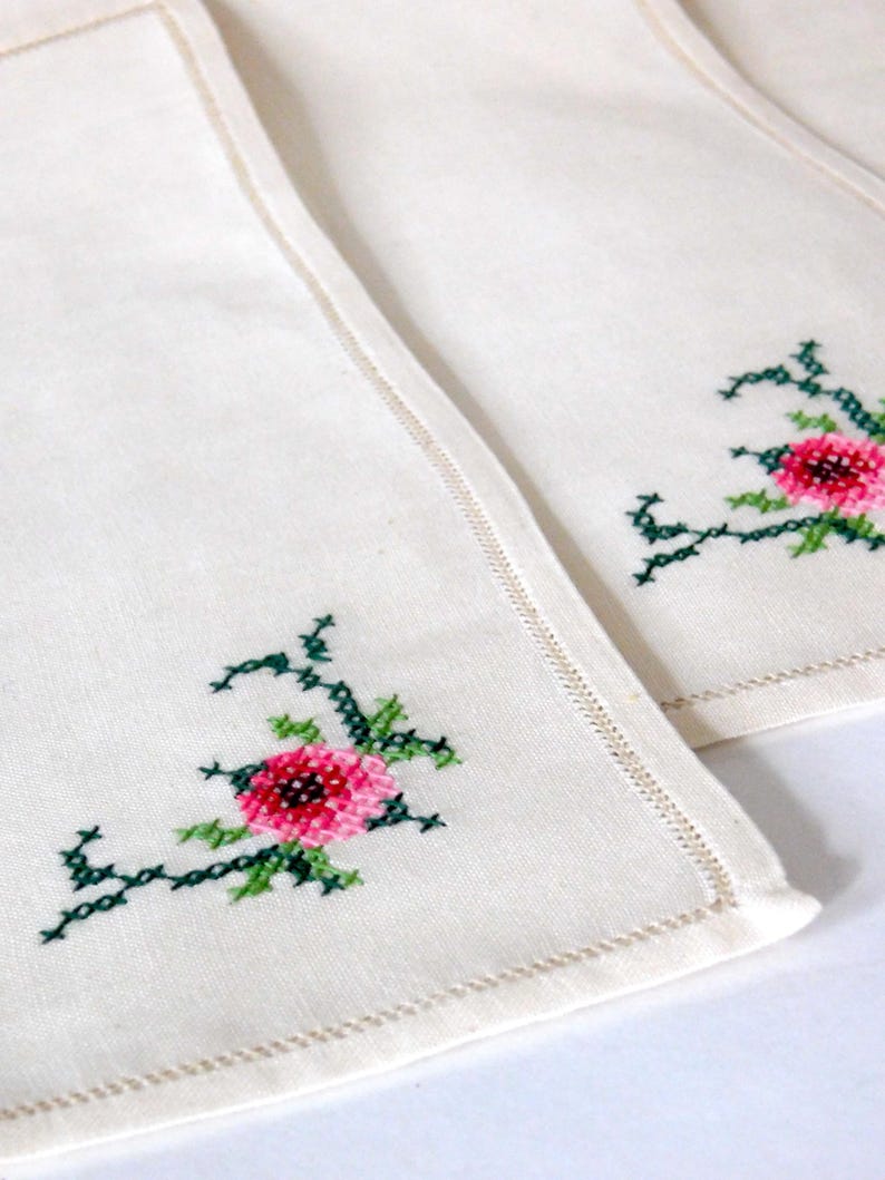 K&ouml;nnte beinhalten: Off-white Leinen-Servietten mit gestickten Blumenmustern. Jede Serviette hat eine kreuzgestickte rosa Blume mit gr&uuml;nen Bl&auml;ttern und Stielen in der Ecke. Die Kanten sind mit einem dekorativen Stich versehen.