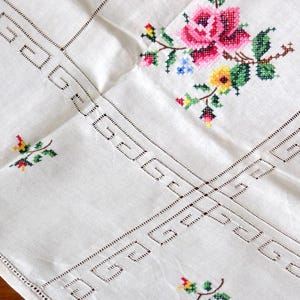 K&ouml;nnte beinhalten: Wei&szlig;es Leinentischtuch mit gestickten floralen Details. Mit einer gro&szlig;en rosa Rose, gr&uuml;nen Bl&auml;ttern und kleinen bunten Blumen. Die R&auml;nder haben ein dekoratives Lochmuster, das dem Textil einen Vintage-Touch verleiht.