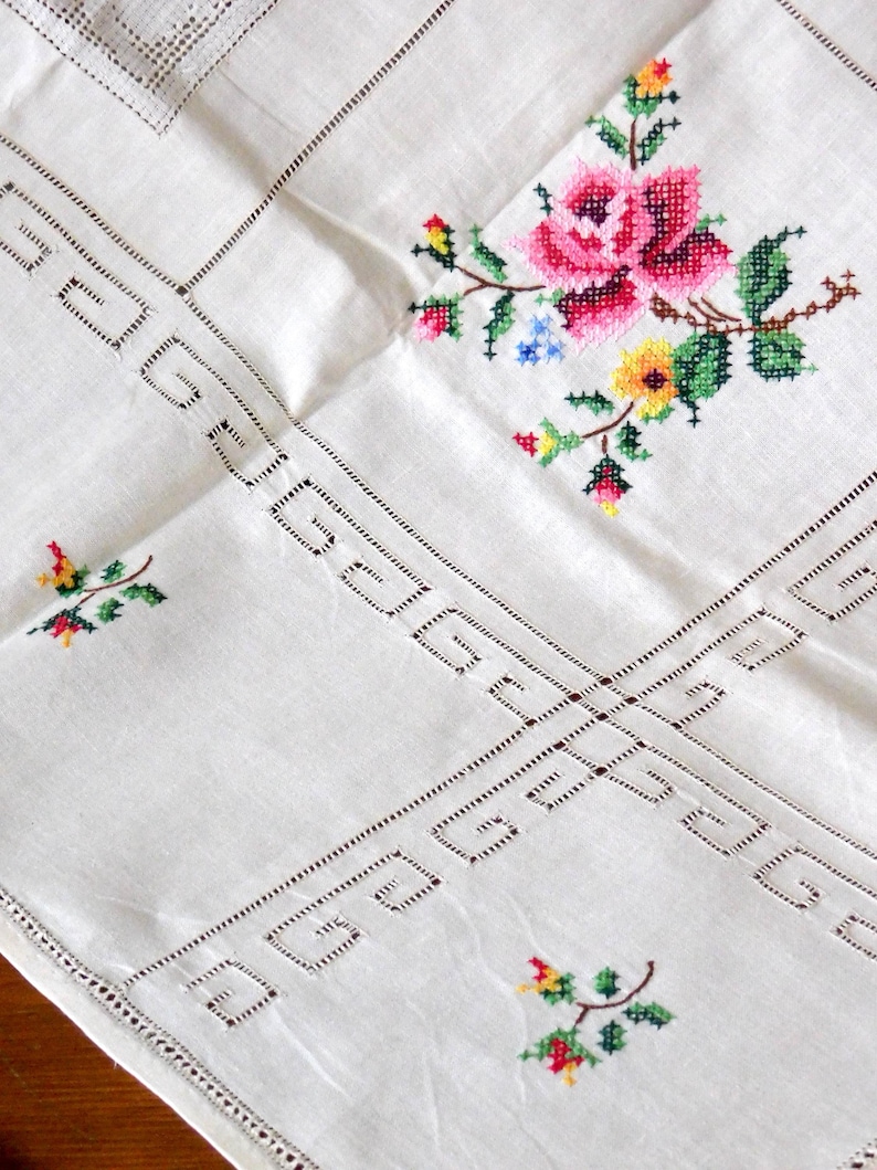 K&ouml;nnte beinhalten: Wei&szlig;es Leinentischtuch mit gestickten floralen Details. Mit einer gro&szlig;en rosa Rose, gr&uuml;nen Bl&auml;ttern und kleinen bunten Blumen. Die R&auml;nder haben ein dekoratives Lochmuster, das dem Textil einen Vintage-Touch verleiht.
