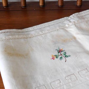 K&ouml;nnte beinhalten: Cremefarbenes Leinentuch mit gesticktem Blumendesign in Rot, Gelb und Gr&uuml;n. Das Tuch hat einen dekorativen Rand mit ausgeschnittenen geometrischen Formen und einen gen&auml;hten Rand. Flecken sind auf dem Stoff sichtbar.