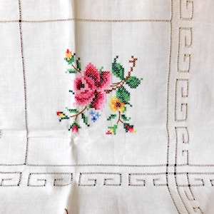 K&ouml;nnte beinhalten: Wei&szlig;es Leinentischtuch mit einem Kreuzstich-Blumenmuster. Das zentrale Motiv zeigt eine rosa Rose mit gr&uuml;nen Bl&auml;ttern und gelben und blauen Akzenten. Der Rand hat ein geometrisches Muster und Lochmusterdetails.