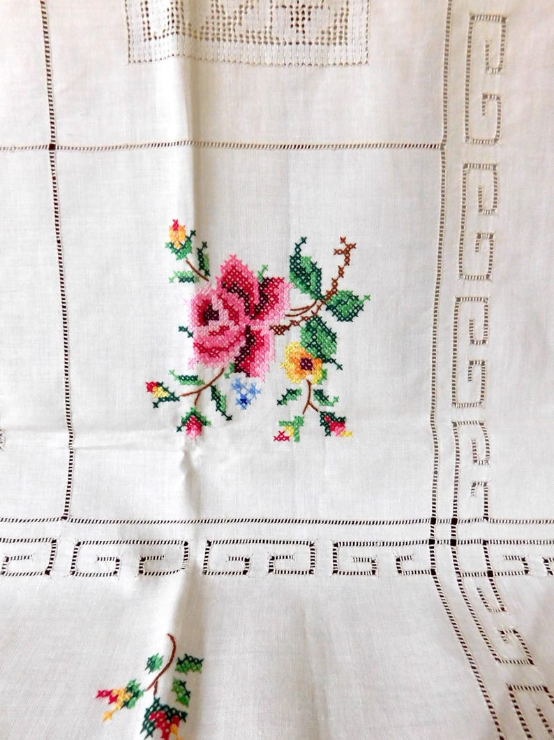K&ouml;nnte beinhalten: Wei&szlig;es Leinentischtuch mit einem Kreuzstich-Blumenmuster. Das zentrale Motiv zeigt eine rosa Rose mit gr&uuml;nen Bl&auml;ttern und gelben und blauen Akzenten. Der Rand hat ein geometrisches Muster und Lochmusterdetails.