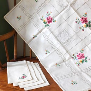 K&ouml;nnte beinhalten: Wei&szlig;es Leinen-Tischdecke und passende Servietten mit gestickten Blumenmustern. Die Tischdecke hat ein Gittermuster mit gestickten Rosen und kleinen floralen Akzenten. Die Servietten haben eine einzelne Blumenstickerei.