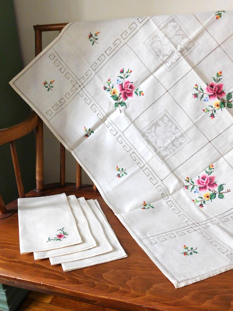 K&ouml;nnte beinhalten: Wei&szlig;es Leinen-Tischdecke und passende Servietten mit gestickten Blumenmustern. Die Tischdecke hat ein Gittermuster mit gestickten Rosen und kleinen floralen Akzenten. Die Servietten haben eine einzelne Blumenstickerei.