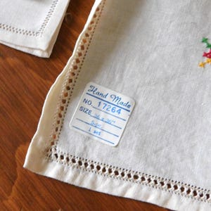 K&ouml;nnte beinhalten: Nahaufnahme eines wei&szlig;en Leinen-Sets mit dekorativen Ausschnittkanten. Das Set enth&auml;lt ein Etikett mit der Aufschrift "Hand Made" und "GR&Ouml;SSE 91 x 91 cm". Das Set hat ein kleines gesticktes Blumenmuster.