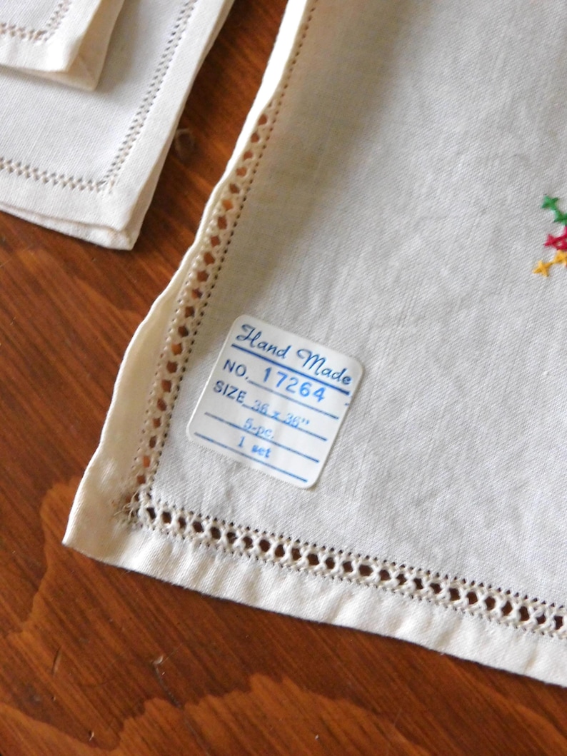K&ouml;nnte beinhalten: Nahaufnahme eines wei&szlig;en Leinen-Sets mit dekorativen Ausschnittkanten. Das Set enth&auml;lt ein Etikett mit der Aufschrift "Hand Made" und "GR&Ouml;SSE 91 x 91 cm". Das Set hat ein kleines gesticktes Blumenmuster.