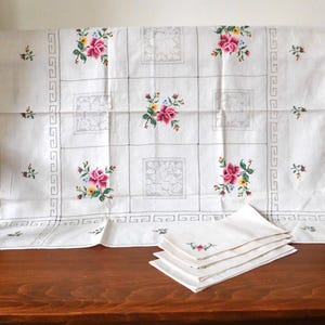 K&ouml;nnte beinhalten: Wei&szlig;es Tischtuch und passende Servietten mit gestickten Blumenmustern. Das Tischtuch hat ein Gittermuster mit gestickten Blumen in Rosa, Gelb und Blau. Die Servietten haben eine &auml;hnliche Blumenstickerei. Ein dekoratives Vintage-Textil.