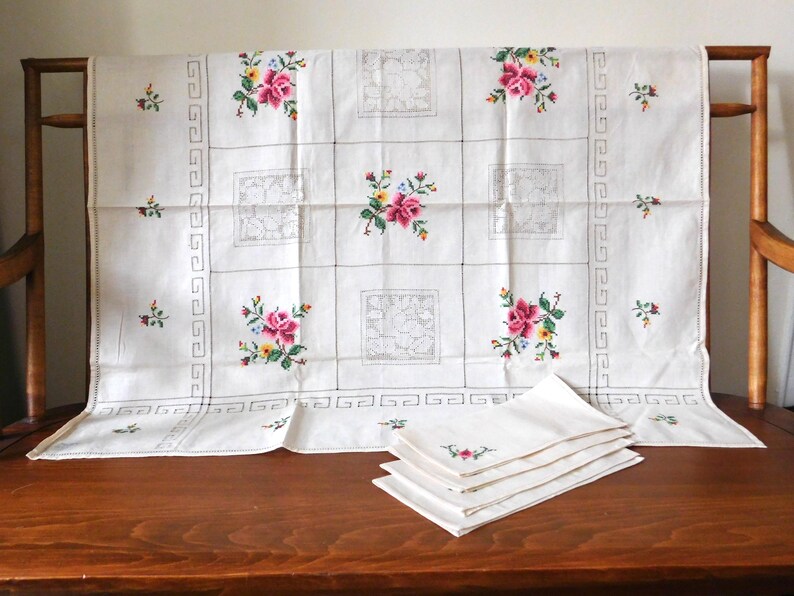 K&ouml;nnte beinhalten: Wei&szlig;es Tischtuch und passende Servietten mit gestickten Blumenmustern. Das Tischtuch hat ein Gittermuster mit gestickten Blumen in Rosa, Gelb und Blau. Die Servietten haben eine &auml;hnliche Blumenstickerei. Ein dekoratives Vintage-Textil.